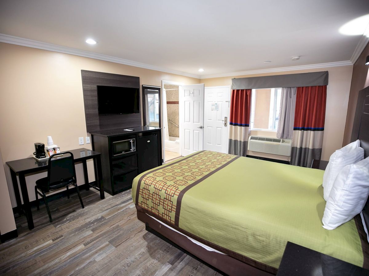 A hotel room with a bed, TV, desk and chair, and a window with curtains. The room has modern decor with neutral colors and wood flooring.