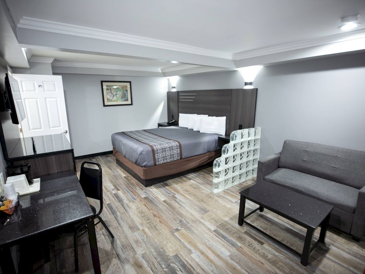 A modern hotel room featuring a bed, grey sofa, desk with chair, and mounted TV, all on wooden flooring, under recessed lighting.