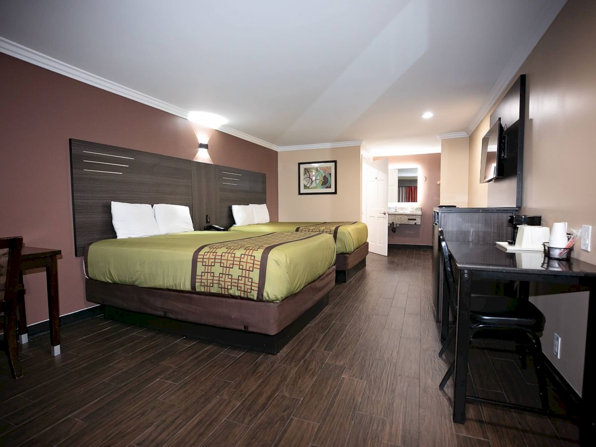 This image shows a hotel room with two double beds, a desk, a TV, and a small dining area. It has wooden flooring and modern furnishings.