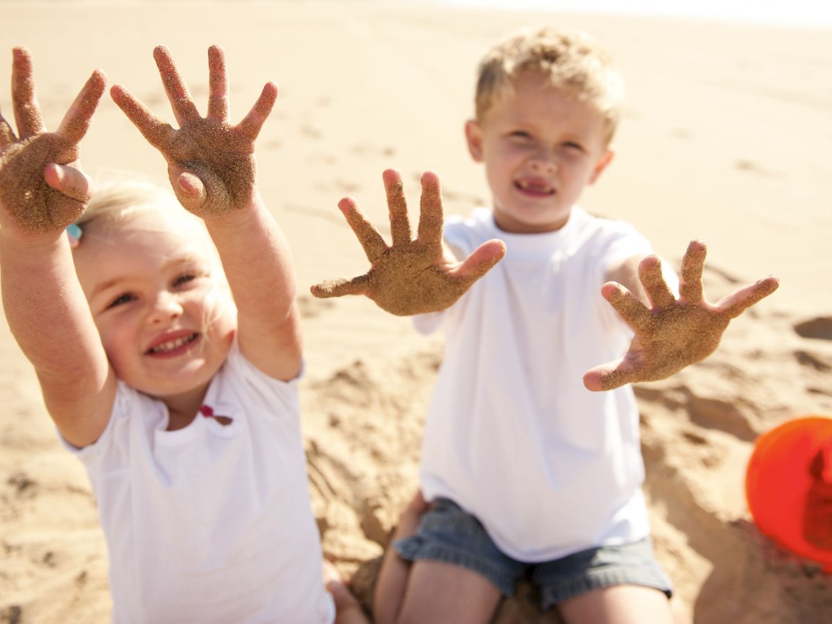Two children are playing on the beach, showing their sandy hands to the camera. There's a red bucket to their right on the sand.