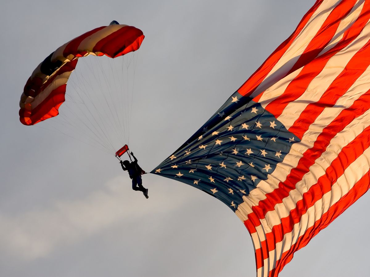 A skydiver is parachuting with a large American flag trailing behind them against a backdrop of a cloudy sky.