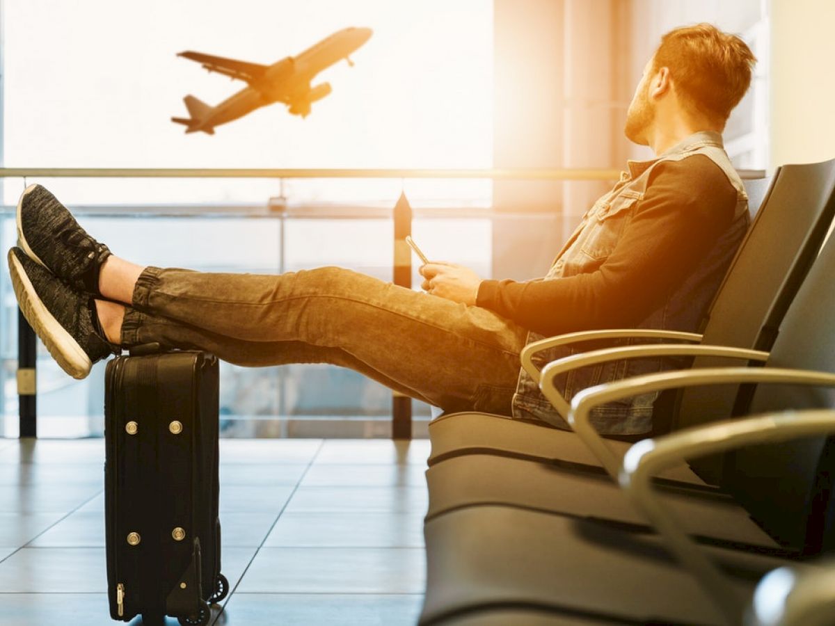 A person is sitting in an airport lounge with feet resting on a suitcase, watching a plane take off through the window.