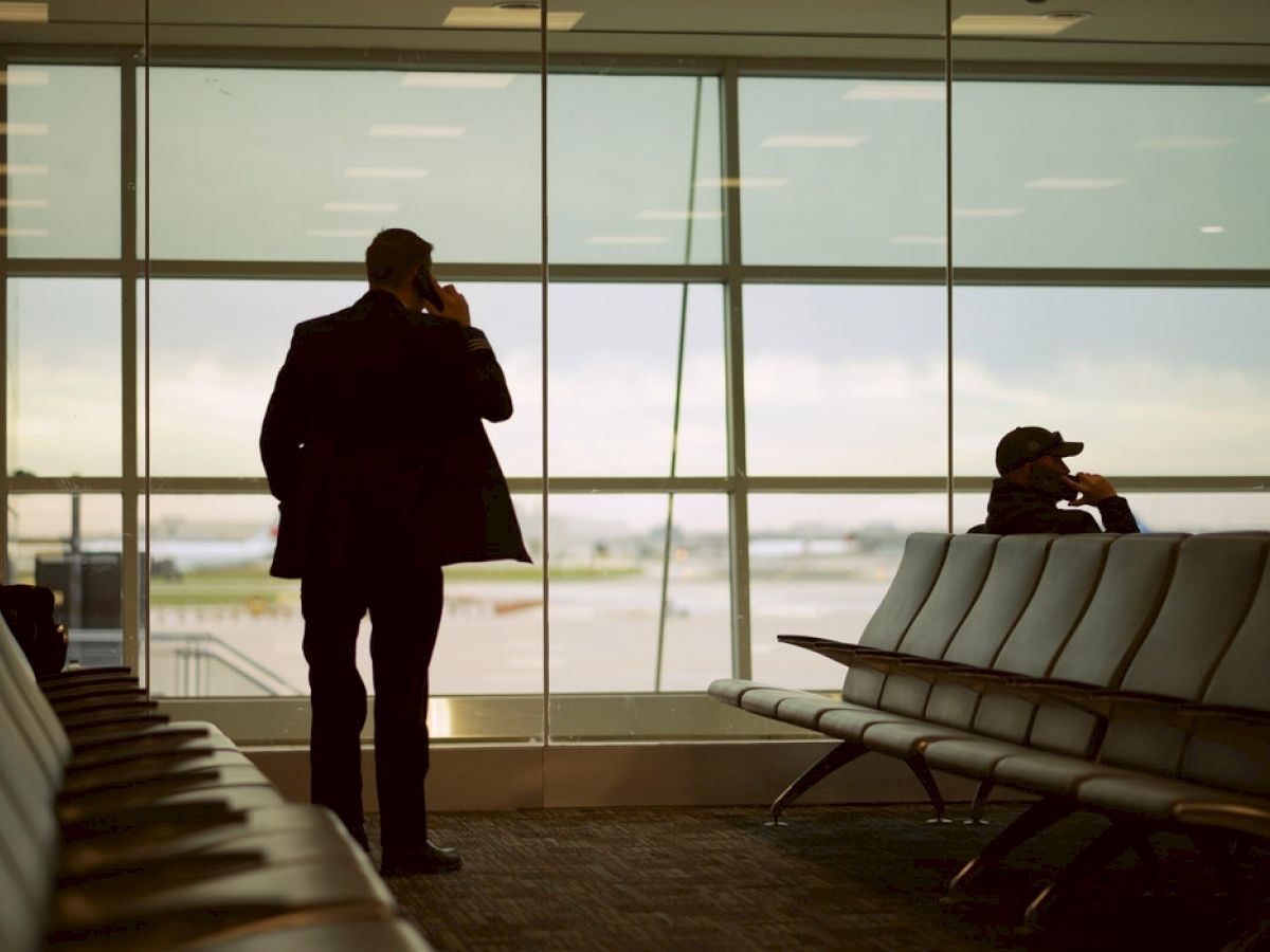 Two people are in an airport waiting area; one is standing and on the phone, while the other is seated near a window with a view of the tarmac.