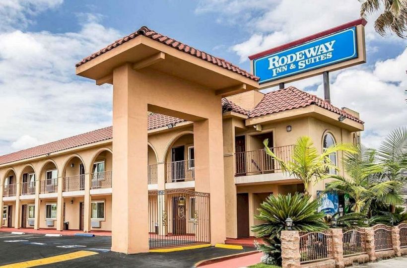 The image shows a tan-colored Rodeway Inn & Suites hotel building with a tiled roof and balconies, surrounded by plants and a clear sky.