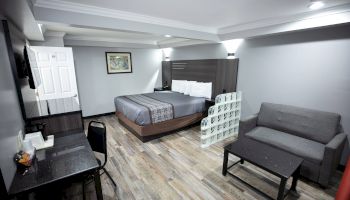 A well-lit hotel room with a bed, sofa, desk, chair, painting, and modern decor. The wooden floor complements the minimalist style.