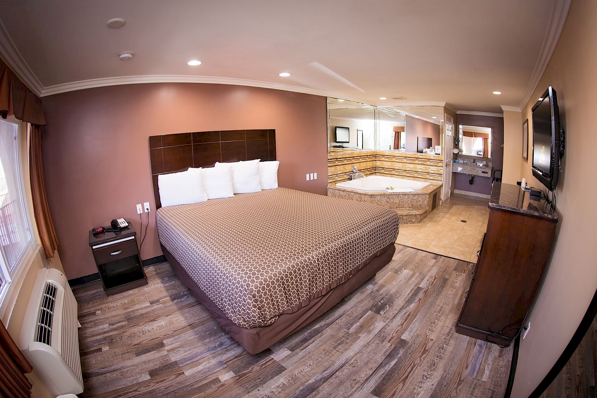 A hotel room with a large bed, bedside table, TV, air conditioner, large mirrors, and a Jacuzzi in the corner, featuring a wooden floor and warm lighting.