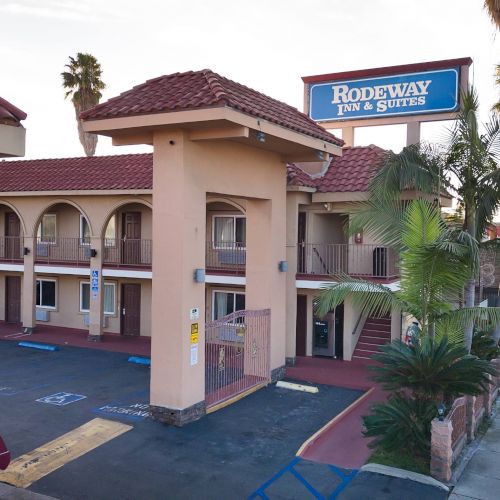 The image shows a Rodeway Inn & Suites motel with two floors, arched balconies, and palm trees, located alongside a street with parked cars.