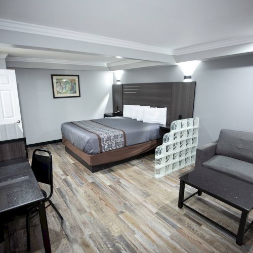 The image shows a modern hotel room with a bed, couch, desk, and art on the wall, featuring minimalist decor and wooden flooring.