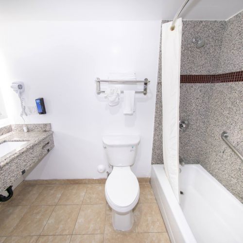 A clean bathroom with a granite countertop, sink, toilet, and a bathtub with a shower curtain; walls are tiled and there are towels hanging.