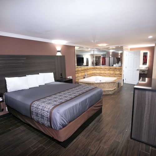 A modern hotel room features a large bed, a jacuzzi, mirrors on the wall, a dresser, and minimalist decor with soft lighting and dark wood flooring.