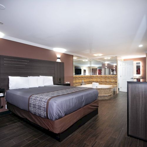The image shows a modern hotel room with a large bed, a TV, a jacuzzi, and a sleek design. The room features dark wood flooring and subdued lighting.