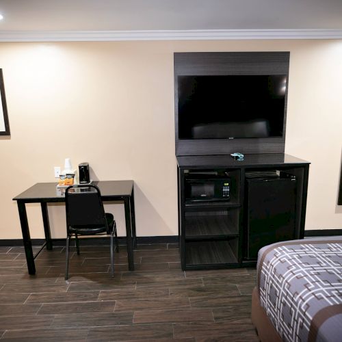 A hotel room features a desk with a coffee maker, a TV mounted on a cabinet, a mirror, a wall painting, and a bed partially in view.