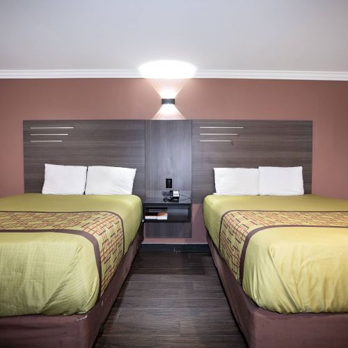 The image shows a hotel room with two double beds, green bedspreads, and a central nightstand under a wall-mounted light. There's art on the right wall.
