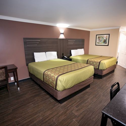 A hotel room features two double beds, a desk, a chair, and a wall-mounted lamp. The room has a dark wood floor and a bathroom in the background.