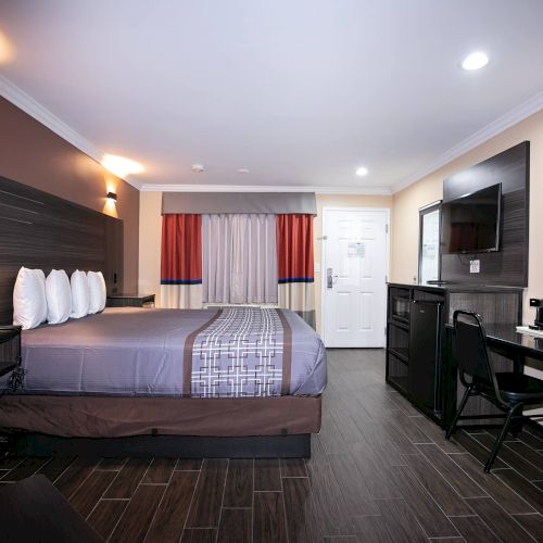 This image shows a modern hotel room with a large bed, a flat-screen TV, a desk and chair, wooden flooring, and a well-lit interior.