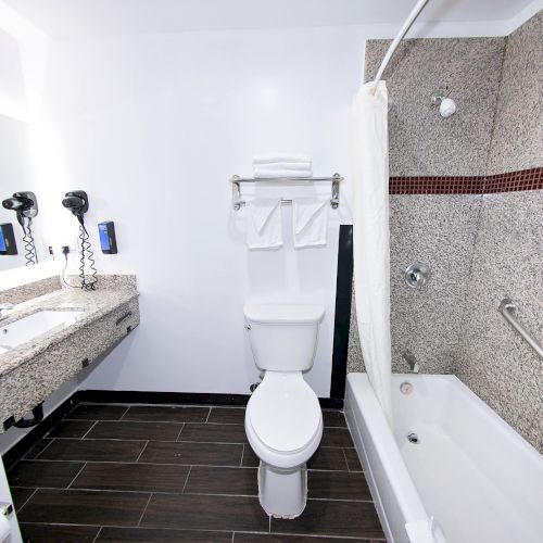 The image shows a clean, modern bathroom with a toilet, sink, mirror, hair dryers, and a bathtub-shower combo featuring a tiled wall.