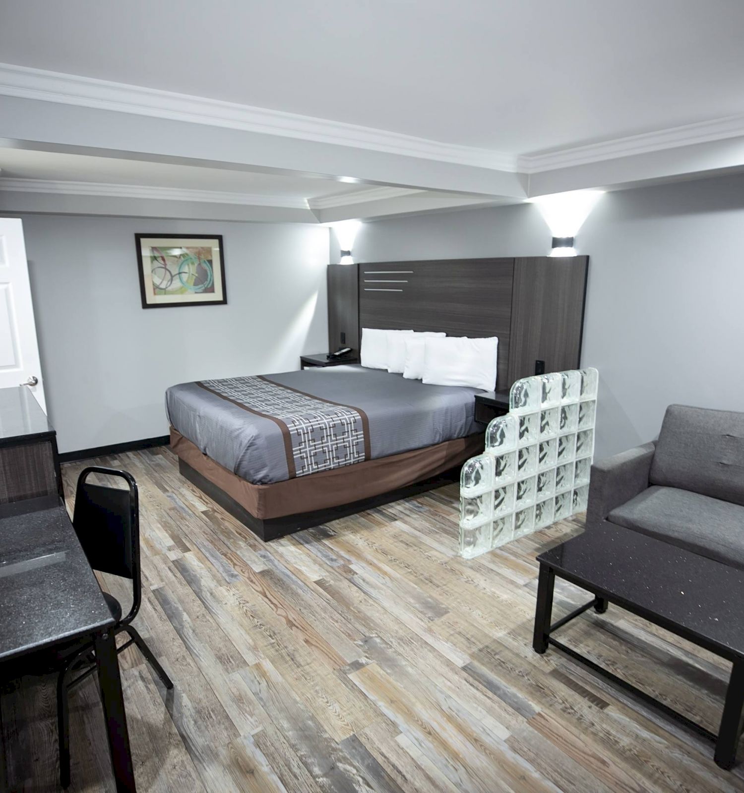 A modern hotel room with a bed, sofa, desk, and TV, featuring wood flooring, a framed picture on the wall, and contemporary furnishings.
