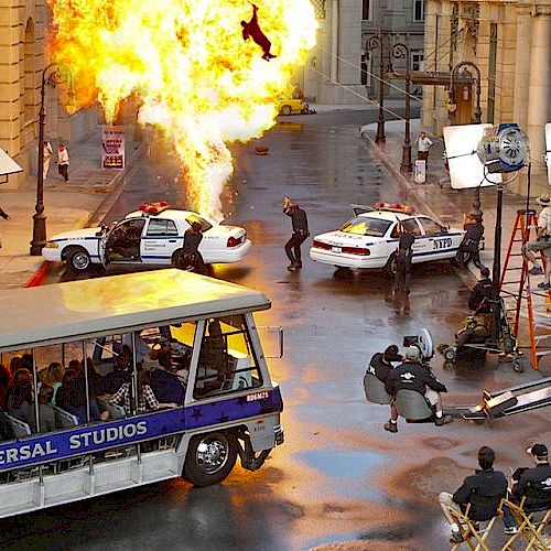 The image shows a film set with a staged explosion, police cars, crew members, lighting equipment, and a Universal Studios tour bus with passengers watching.