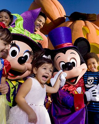 A group of children in costumes are happily interacting with two mascots dressed as Halloween-themed characters, in front of a pumpkin backdrop.