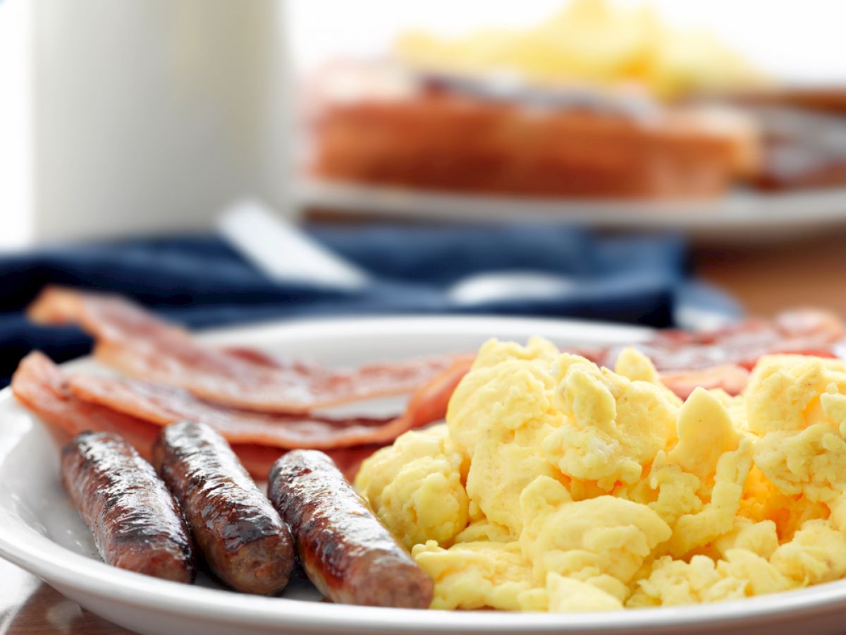 The image shows a plate of breakfast food including scrambled eggs, sausages, and bacon, with toast and a beverage in the background.